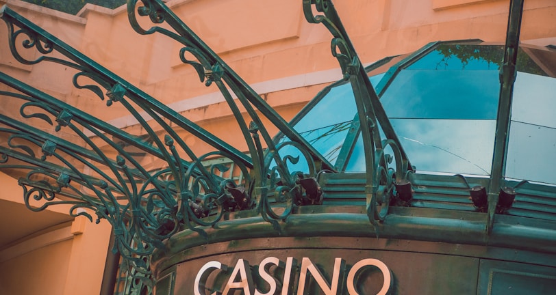 The image displays the entrance to a casino, characterized by detailed and ornate architectural elements with green metal structures and a sign that reads 'CASINO'. The background includes a glass dome and a peach-colored wall.