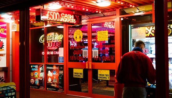 A brightly lit entrance to a casino with neon signs, including one advertising a £500 jackpot. The doors have clear signage indicating entry is restricted to those over 18. A person in a red jacket stands to the right, possibly interacting with a machine or display.