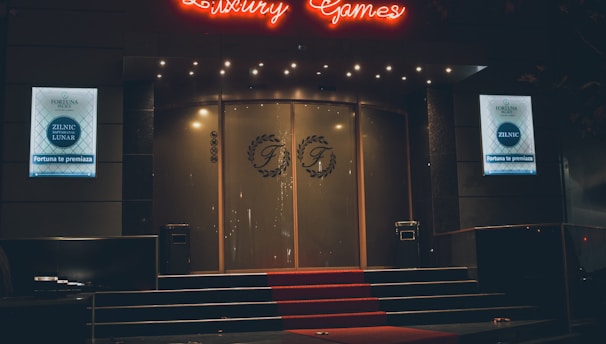 A neon-lit casino entrance with a red carpet leading up to large double doors. The sign reads 'Fortuna Palace Luxury Games' in bright red and white neon lights. The dimly lit atmosphere suggests a night-time setting. There are two digital displays on either side of the entrance showing promotional content.