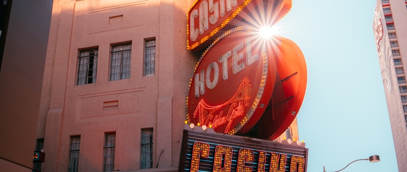 A vibrant city scene featuring a tall, illuminated casino sign with retro-style neon lights. The sign dominates the building facade with the words 'CASINO' and 'HOTEL'. Sunlight peeks through, creating a dramatic lens flare effect. The street is lively, with pedestrians casually walking and a hint of urban hustle.
